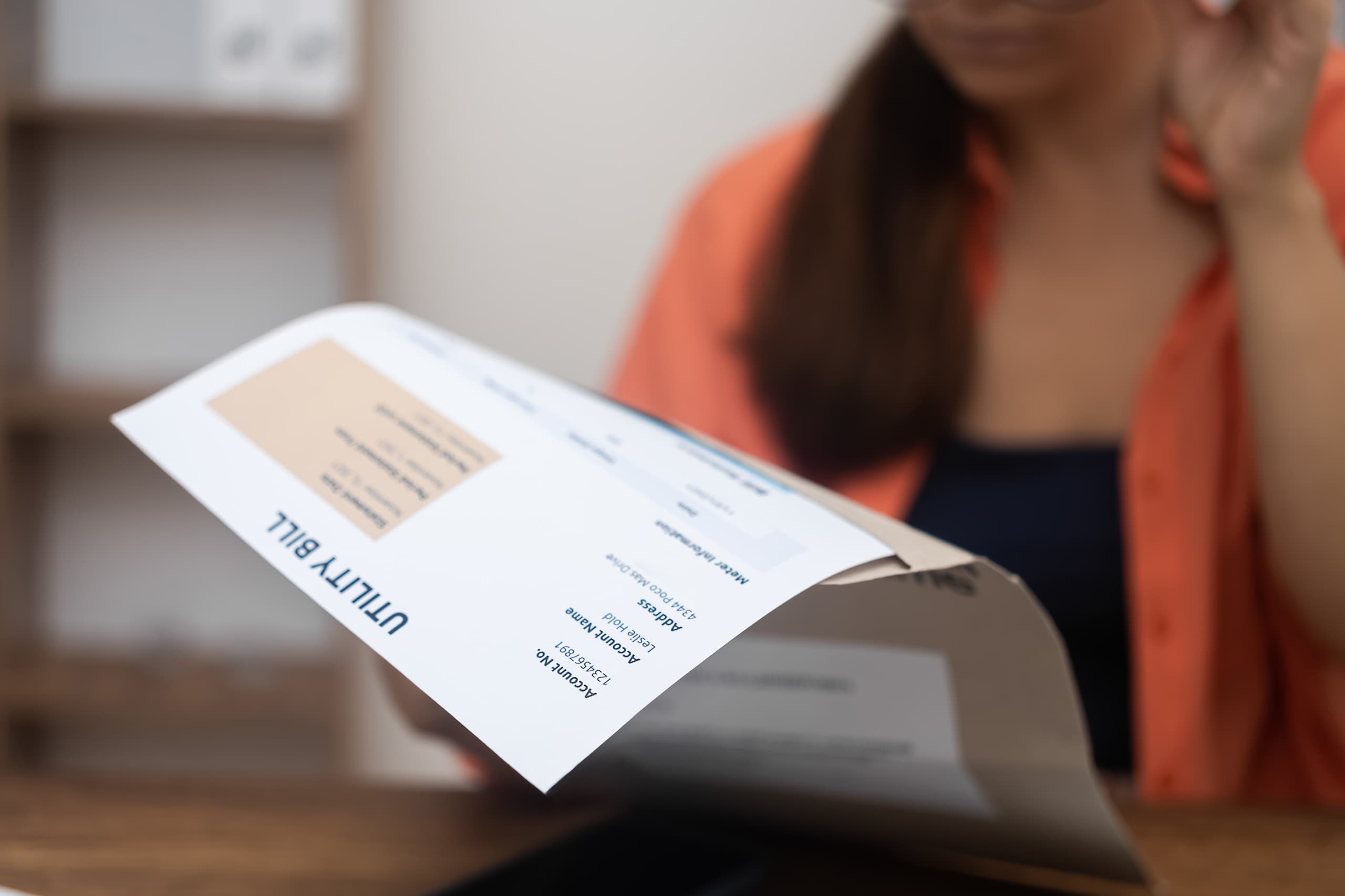 A close-up of a woman in an orange cardigan holding and reading a utility bill, with account name and address details visible on the document, sitting at a desk with a blurred office background.
